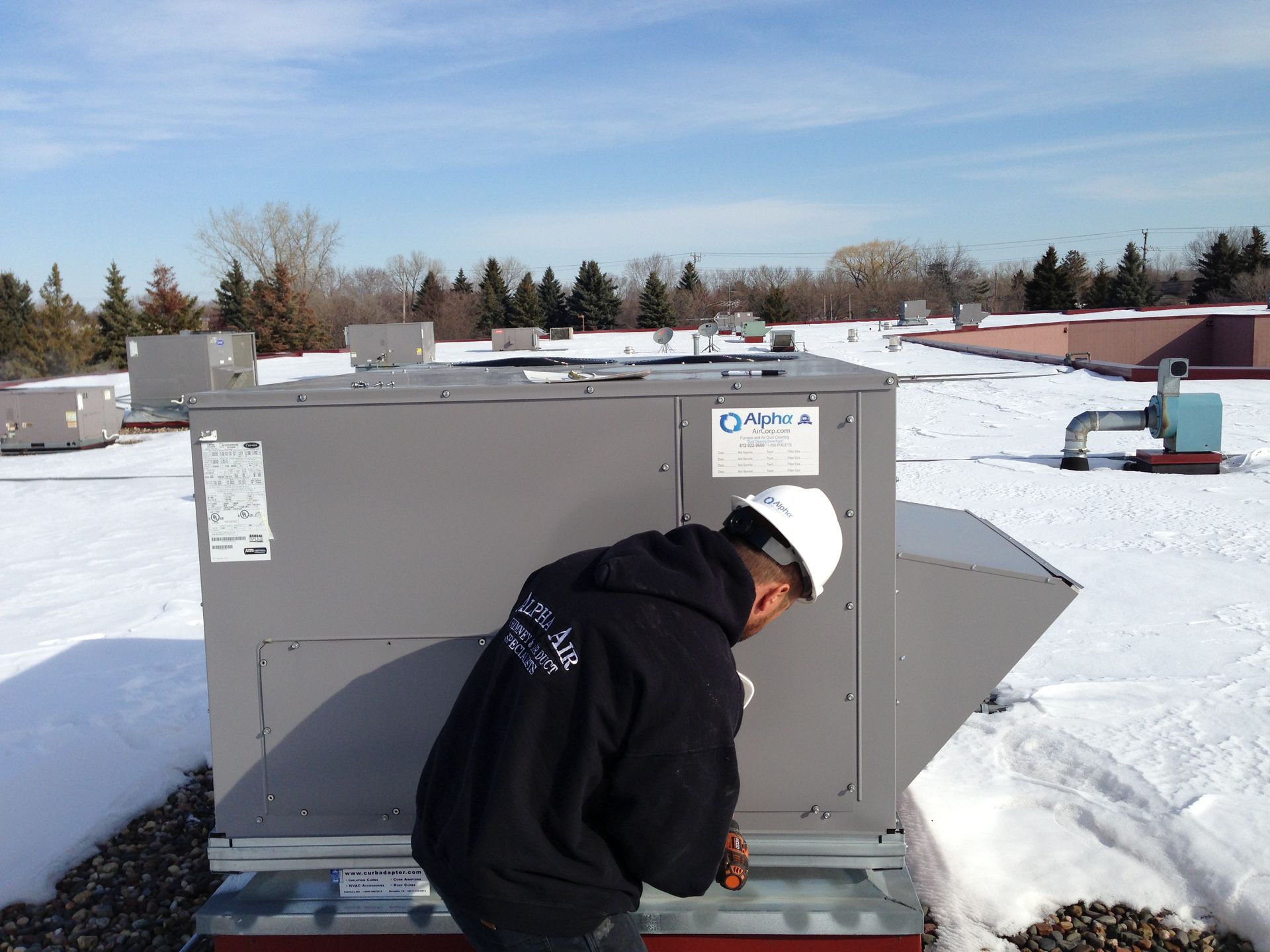 Person in a hard hat working on an HVAC unit on a snowy rooftop.