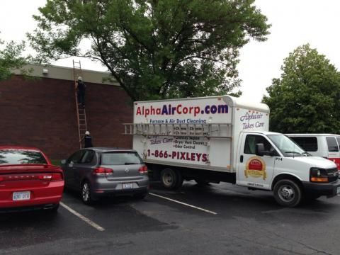A work truck, Alpha Air Corp, parked near a building. Two people on a ladder, working on the building's exterior.