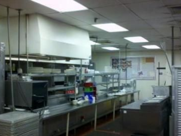 Commercial kitchen with stainless steel counters, range hood, and overhead lighting.