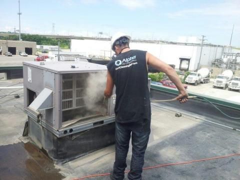 Man cleaning HVAC unit on a rooftop. White vapor is visible. The man wears a hard hat and Alpha-branded shirt.