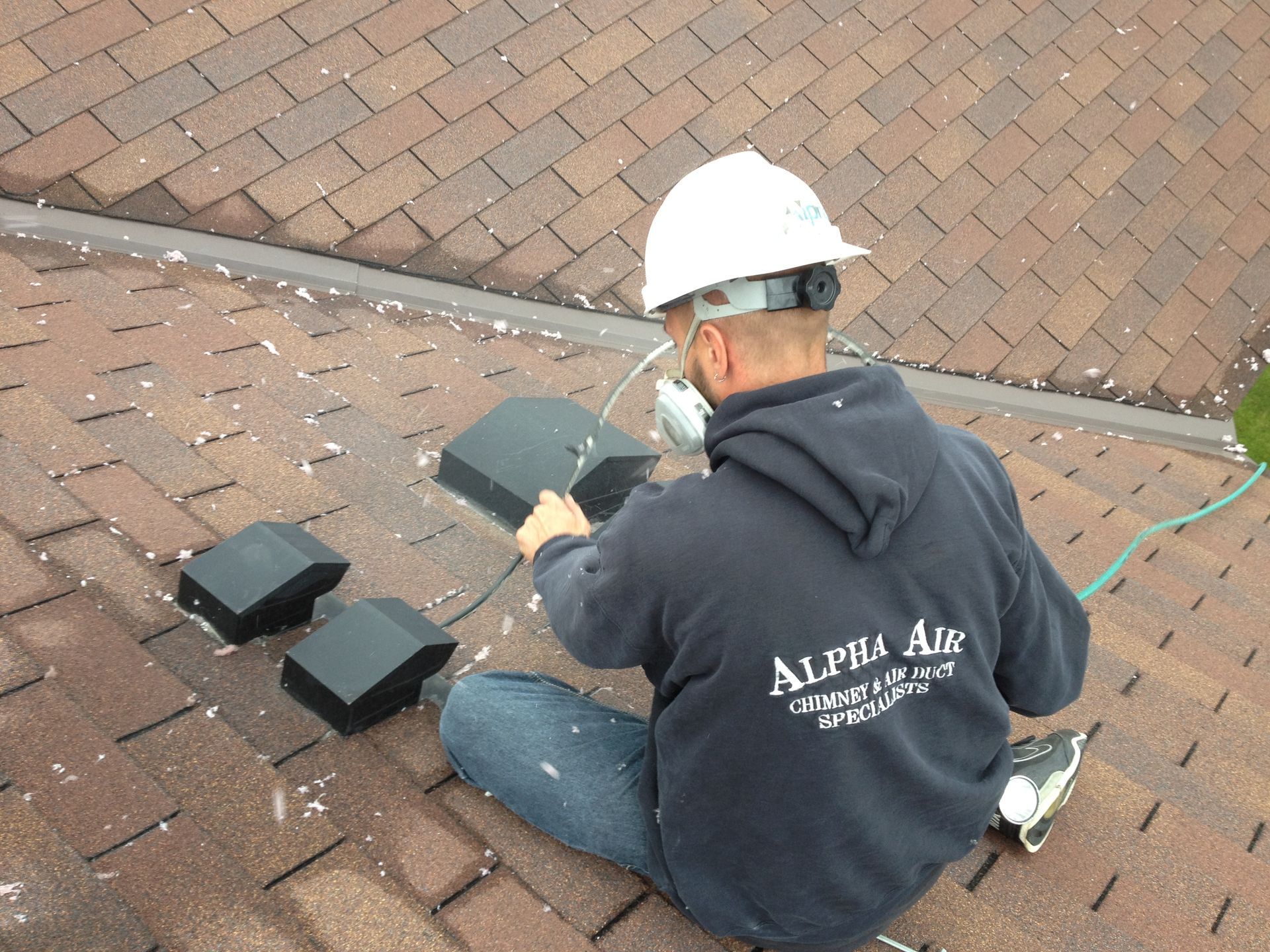 Roofer on a brown shingled roof installing black vents, wearing a hard hat and Alpha Air sweatshirt.