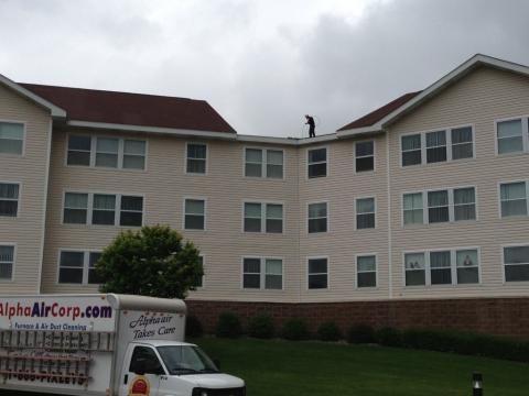 A worker on a roof of a multi-story building, an Alpha Air Corp. van parked on the lawn below.