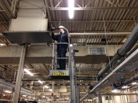 Man on a lift working on an industrial ceiling duct. Gray, metallic, interior setting.