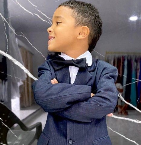 Niño pequeño con traje y pajarita, con los brazos cruzados, sonriendo.