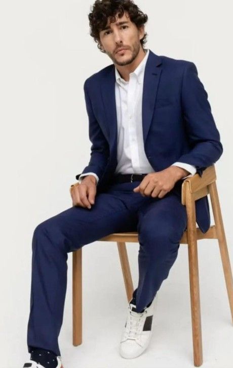 Hombre con traje azul, camisa blanca y zapatillas deportivas, sentado en una silla de madera sobre un fondo blanco.