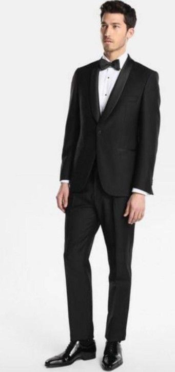 Hombre con esmoquin negro, pajarita negra, camisa blanca y zapatos negros brillantes, sobre fondo blanco.