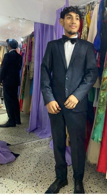 Hombre con traje oscuro y pajarita, de pie en una tienda con percheros y un espejo.