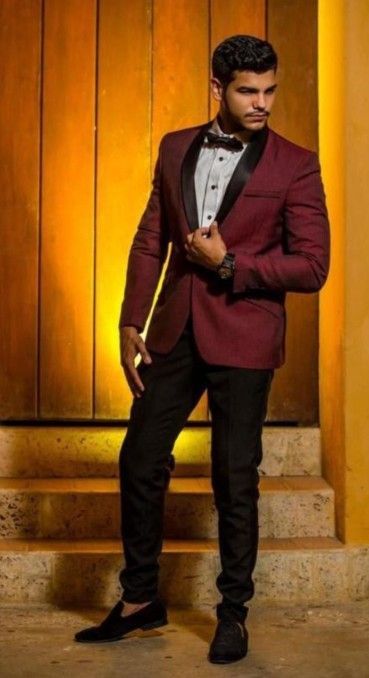 Un hombre con traje burdeos, pantalones negros y pajarita está de pie en unos escalones.