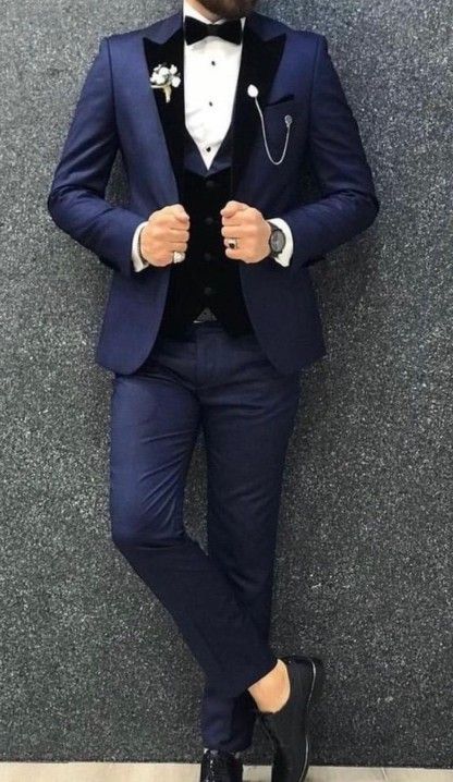 Hombre con traje azul marino, solapas y chaleco negros, pajarita y reloj de bolsillo; de pie frente a una pared texturizada.