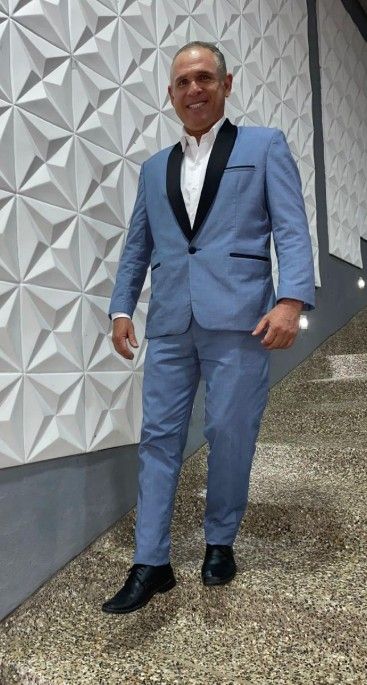 Un hombre con traje azul, solapas y zapatos negros, sonriente, está de pie frente a una pared geométrica blanca.