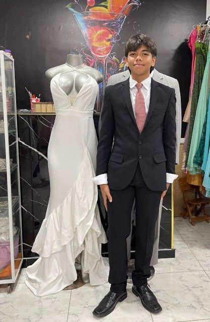 Joven con traje de pie junto a un vestido blanco en una tienda de ropa.