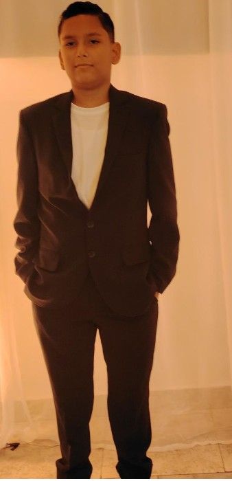 Una persona vestida con traje negro, con las manos en los bolsillos, está de pie contra un fondo claro.