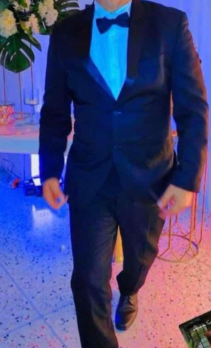 Hombre con esmoquin negro y camisa azul, con pajarita, de pie en el interior con decoración de fiesta.