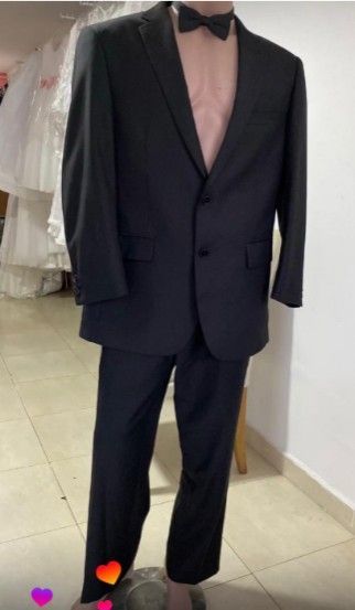 Traje negro con pajarita en maniquí. Interior, contra pared blanca y vestidos.