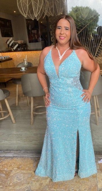 Mujer con un vestido formal azul claro de lentejuelas, de pie junto a una pared de cristal, sonriendo, con un collar.