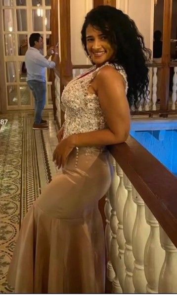 Mujer con vestido de gala se apoya en una barandilla en el interior; vestido beige y blanco, cabello oscuro rizado.