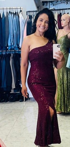 Una mujer con un vestido burdeos de lentejuelas de un solo hombro posa, sosteniendo un teléfono, en una tienda.