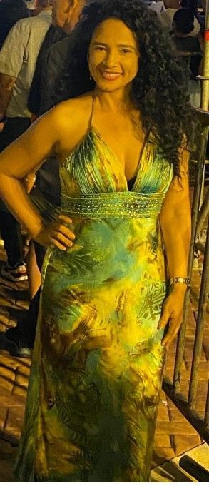 Una mujer con un vestido verde y dorado sonríe, posando con la mano en la cadera. Al fondo se ven personas y una barandilla.