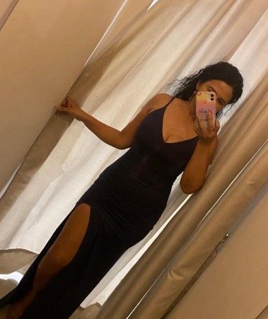 Mujer con un vestido oscuro sin mangas y una abertura hasta el muslo, tomándose una selfie en una habitación