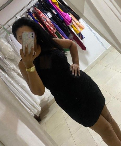 Mujer con vestido negro tomándose una selfie frente al espejo en una tienda de ropa, con la mano en la cadera.