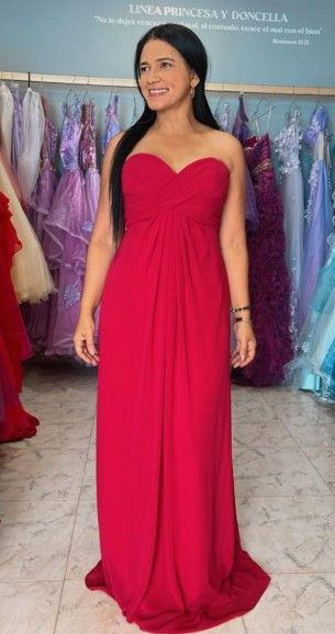 Una mujer con un vestido rojo sin tirantes sonríe, de pie en una tienda de vestidos. Al fondo, se ven vestidos colgados.