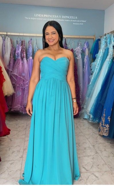 Una mujer con un vestido turquesa sin tirantes sonríe en una tienda de vestidos. Los vestidos adornan la pared.