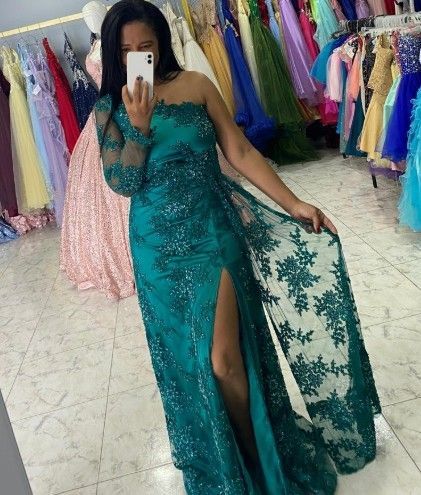 Mujer con vestido azul verdoso de un solo hombro con abertura, sosteniendo un teléfono en una tienda de vestidos.