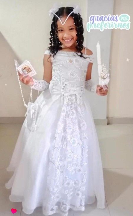 Niña pequeña con vestido blanco de comunión, sonriendo, sosteniendo una vela y un pequeño libro