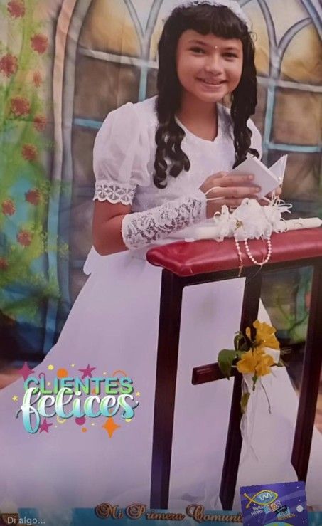 Niña vestida de blanco en su Primera Comunión. Sostiene un papel, sonriendo. Mesa roja con flores.