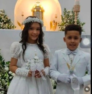 Dos niños vestidos de etiqueta blanca, sosteniendo velas, frente a un altar.