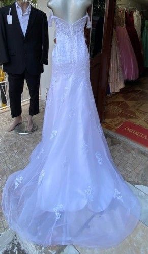 Vestido de novia blanco con hombros descubiertos y larga cola, expuesto en el escaparate de una tienda junto a un traje.