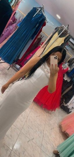 Una mujer con vestido blanco se toma una selfie frente al espejo en una tienda de vestidos. Un vestido rojo cuelga cerca.
