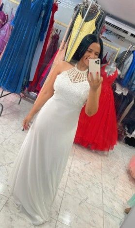Mujer con vestido blanco de cuello halter, sosteniendo un teléfono, tomándose una selfie en una tienda de vestidos.