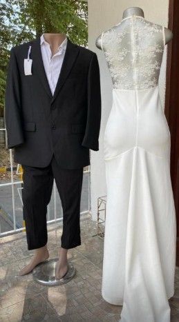 Maniquí con traje negro junto a un vestido de novia blanco sobre un maniquí; al aire libre.