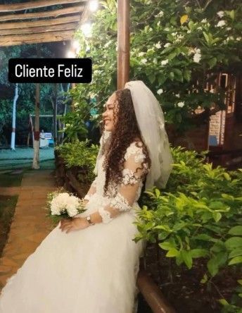 La novia, vestida con un traje blanco y velo, sonríe sosteniendo un ramo, en un entorno al aire libre rodeado de vegetación.