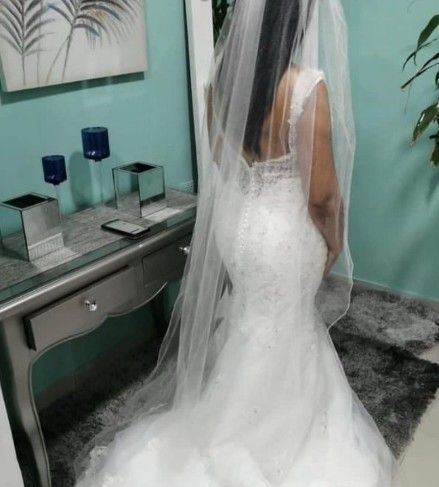 Novia con vestido blanco de corte sirena y velo largo. De pie en el interior, cerca de una mesa.