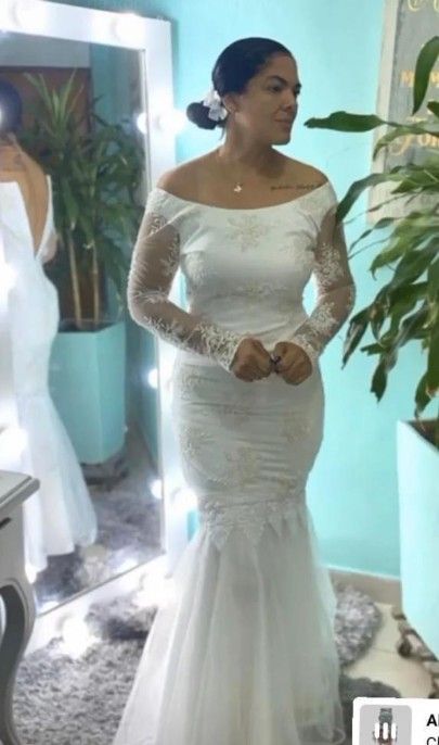 Mujer con vestido de novia blanco de manga larga y hombros descubiertos, mirando hacia otro lado, frente a un espejo.
