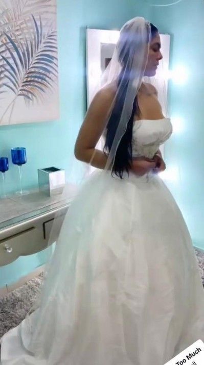 Mujer con vestido de novia blanco sin tirantes y con velo, de pie en una habitación con pared azul claro.
