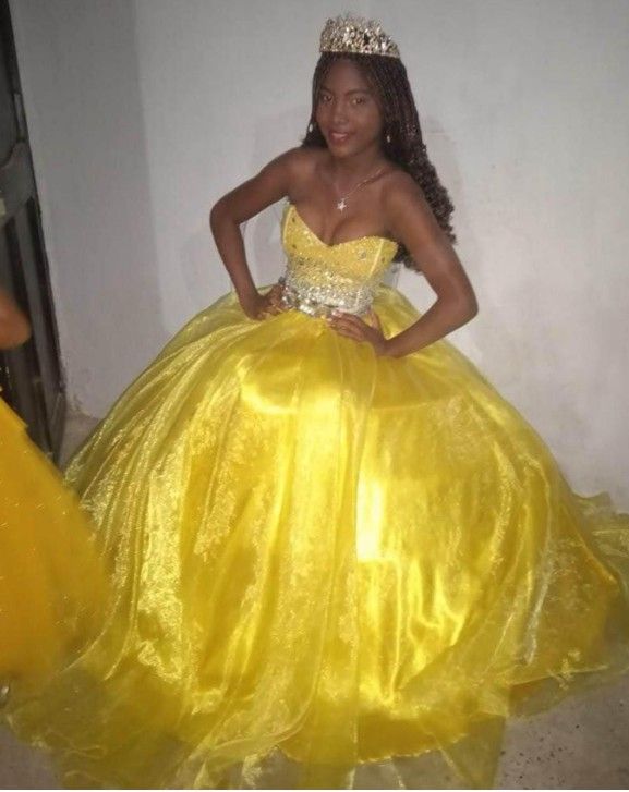 Mujer con vestido de gala amarillo y tiara, posando con las manos en las caderas.