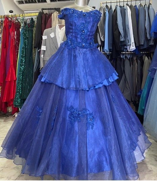 Vestido de gala azul con escote de hombros descubiertos, falda escalonada y adornos florales, exhibido en una tienda de ropa.