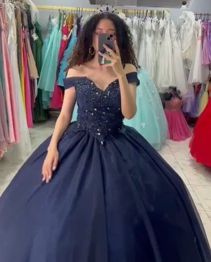 Mujer con vestido de gala azul marino con hombros descubiertos, tomándose selfie frente al espejo en una tienda de vestidos