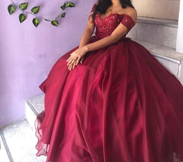mujer con vestido de gala color burdeos con hombros descubiertos, sentada en unos escalones blancos junto a una pared lila