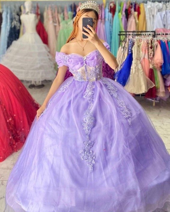 Mujer con un vestido de gala color lavanda, posando con un teléfono. Vestidos de varios colores cuelgan al fondo.