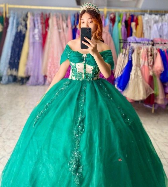 Mujer con vestido de gala verde esmeralda, sosteniendo el teléfono para tomarse una selfie, de pie frente a otros vestidos.