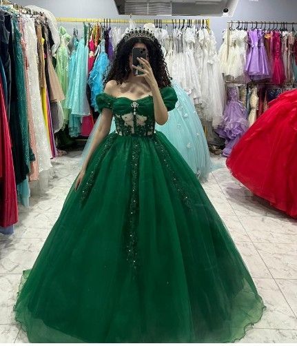 Mujer con vestido de gala verde, sosteniendo un teléfono, dentro de una tienda de vestidos con estantes repletos de vestidos