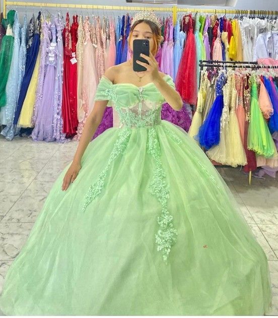 Una mujer con vestido de gala verde claro posa frente a percheros llenos de vestidos coloridos, tomarse una selfie.