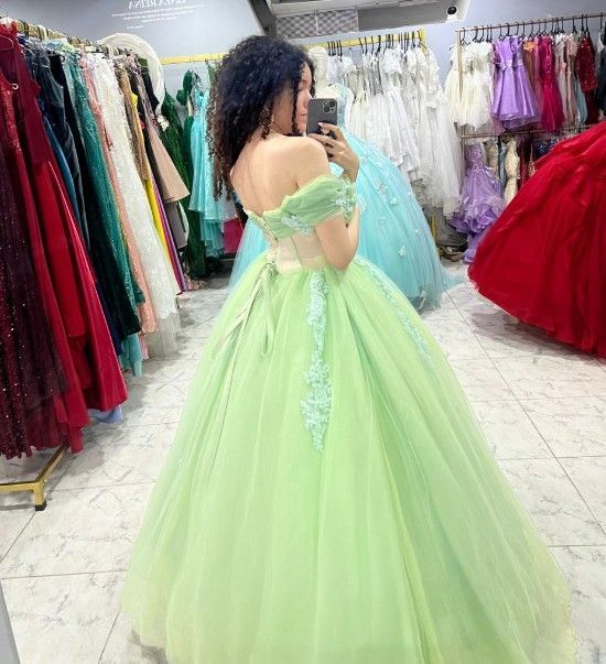 Mujer con un vestido de gala verde claro posa en una tienda de vestidos, tomándose una selfie en el espejo.