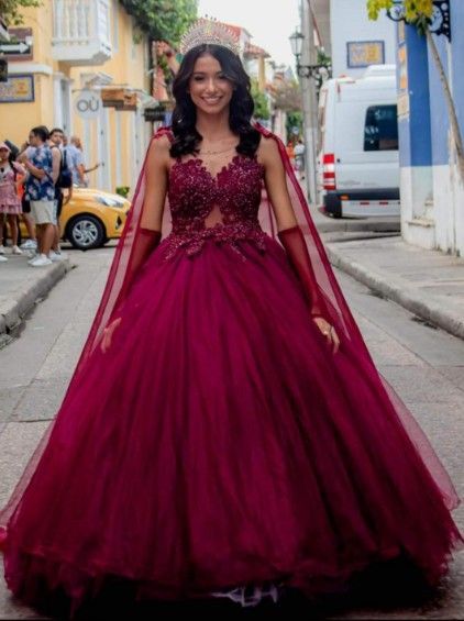 Mujer con vestido de gala color burdeos y capa posando en una calle empedrada.