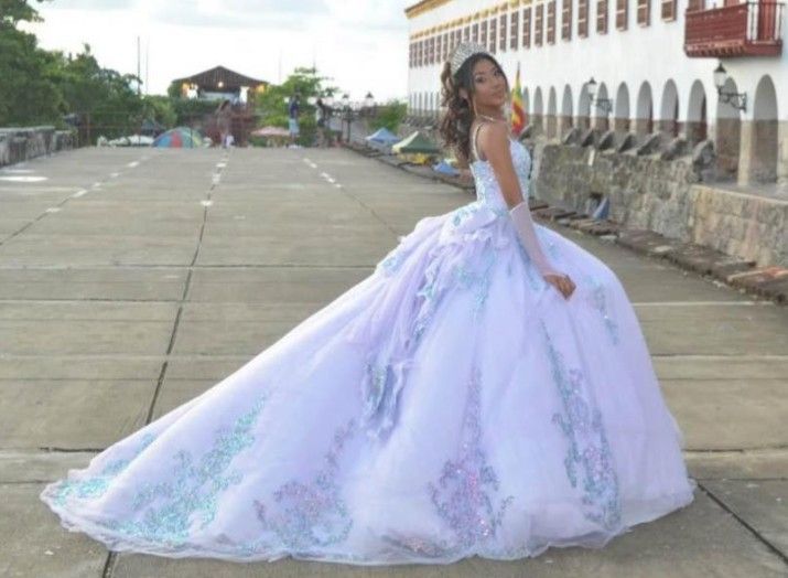 Mujer con vestido de gala color lavanda posa sobre una acera de concreto, con la cola del vestido ondeando