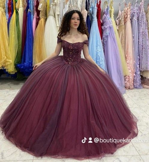 Mujer con un vestido de gala color burdeos, con los hombros al descubierto, posando delante de vestidos coloridos.